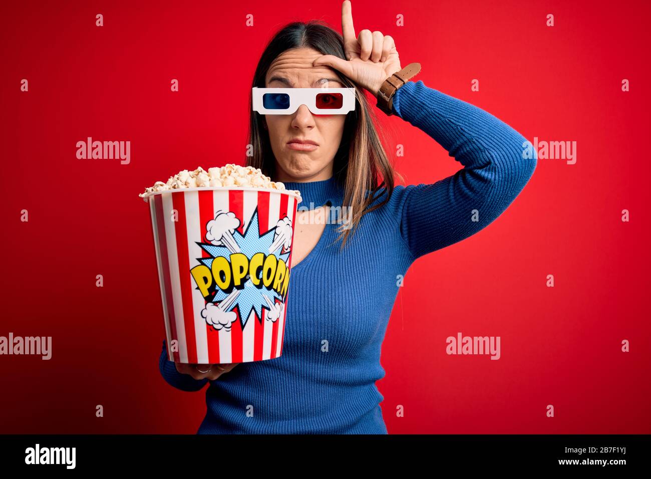 Joven mujer rubia con gafas 3d y comiendo un paquete de palomitas viendo  una película en el cine haciendo diversión de la gente con los dedos en la  frente haciendo perdedor Fotografía