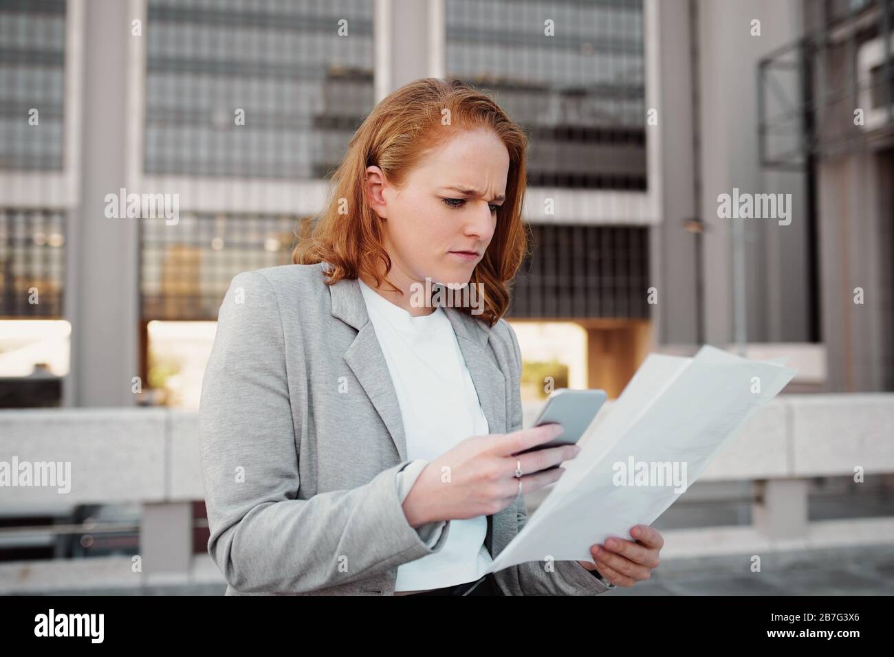 Mujer frustrada con ropa formal utilizando el smartphone mientras lee documentos y realiza cambios al aire libre Foto de stock