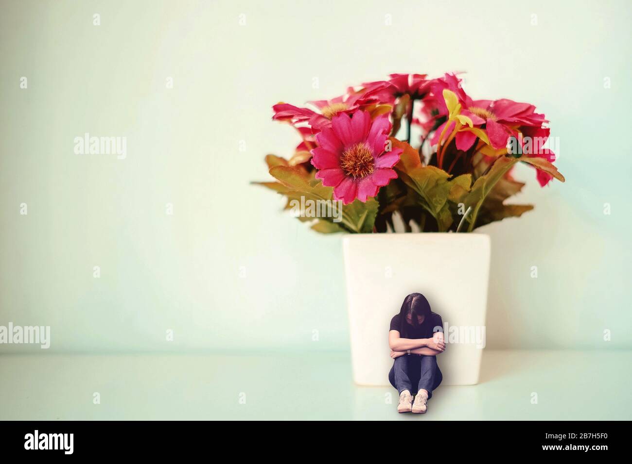 Triste mujer deprimida sentada con la espalda se inclina contra la olla blanca con flor roja falsa. Concepto mínimo de tristeza, soledad. Foto de stock