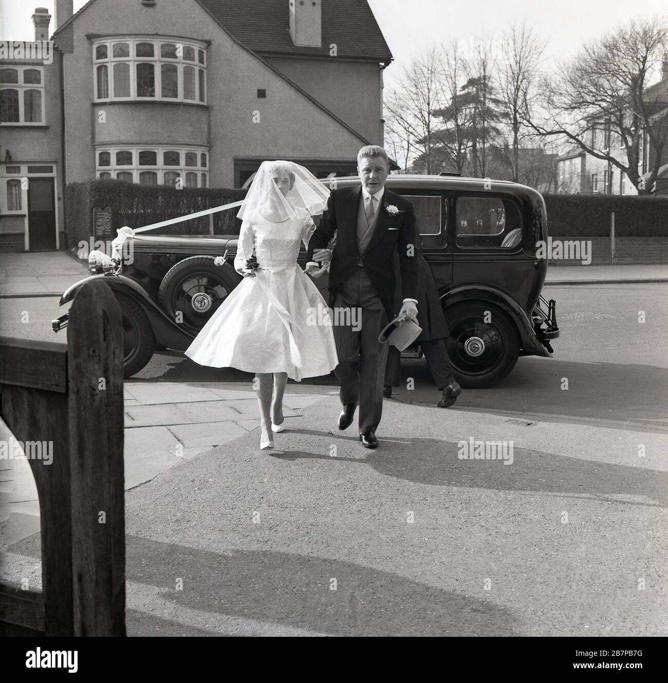 1960, histórico, en el exterior en una calle suburbana, un coche de época aparcado en la carretera, mientras una novia llega para su boda, sureste de Londres, Inglaterra, Reino Unido. Foto de stock