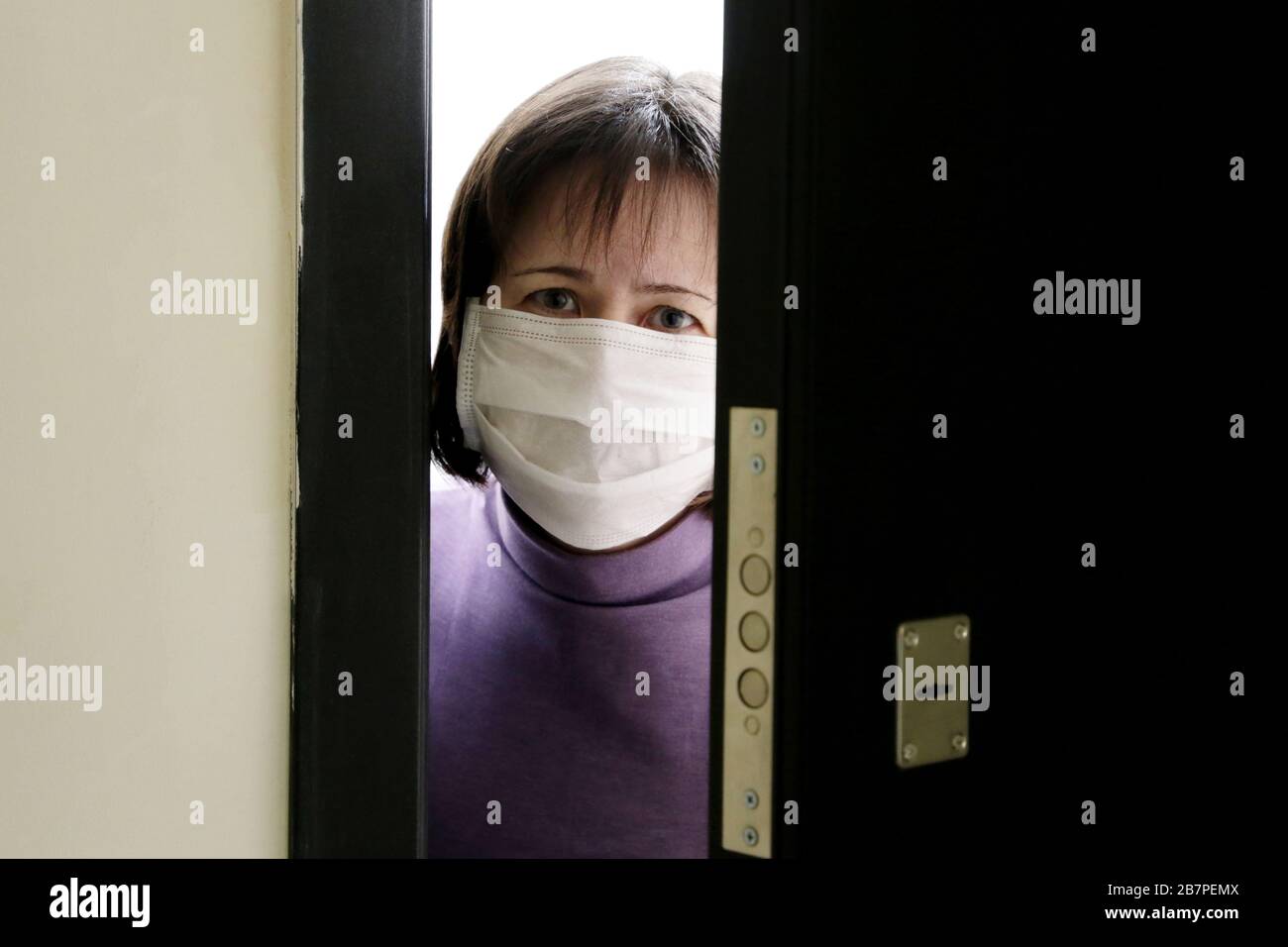 Cuarentena domiciliaria durante la epidemia de coronavirus COVID-19. Mujer preocupada en una máscara médica de pie en la puerta abierta Foto de stock