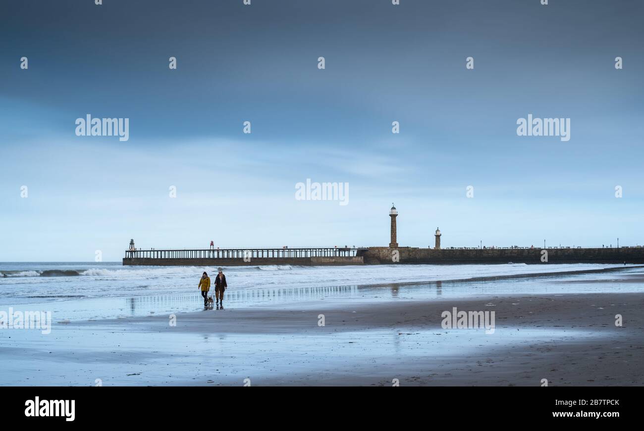 Dos personas caminan a sus perros en la playa de Whitby, con el muelle Whitby al fondo, la costa de North Yorkshire, Inglaterra, Reino Unido Foto de stock