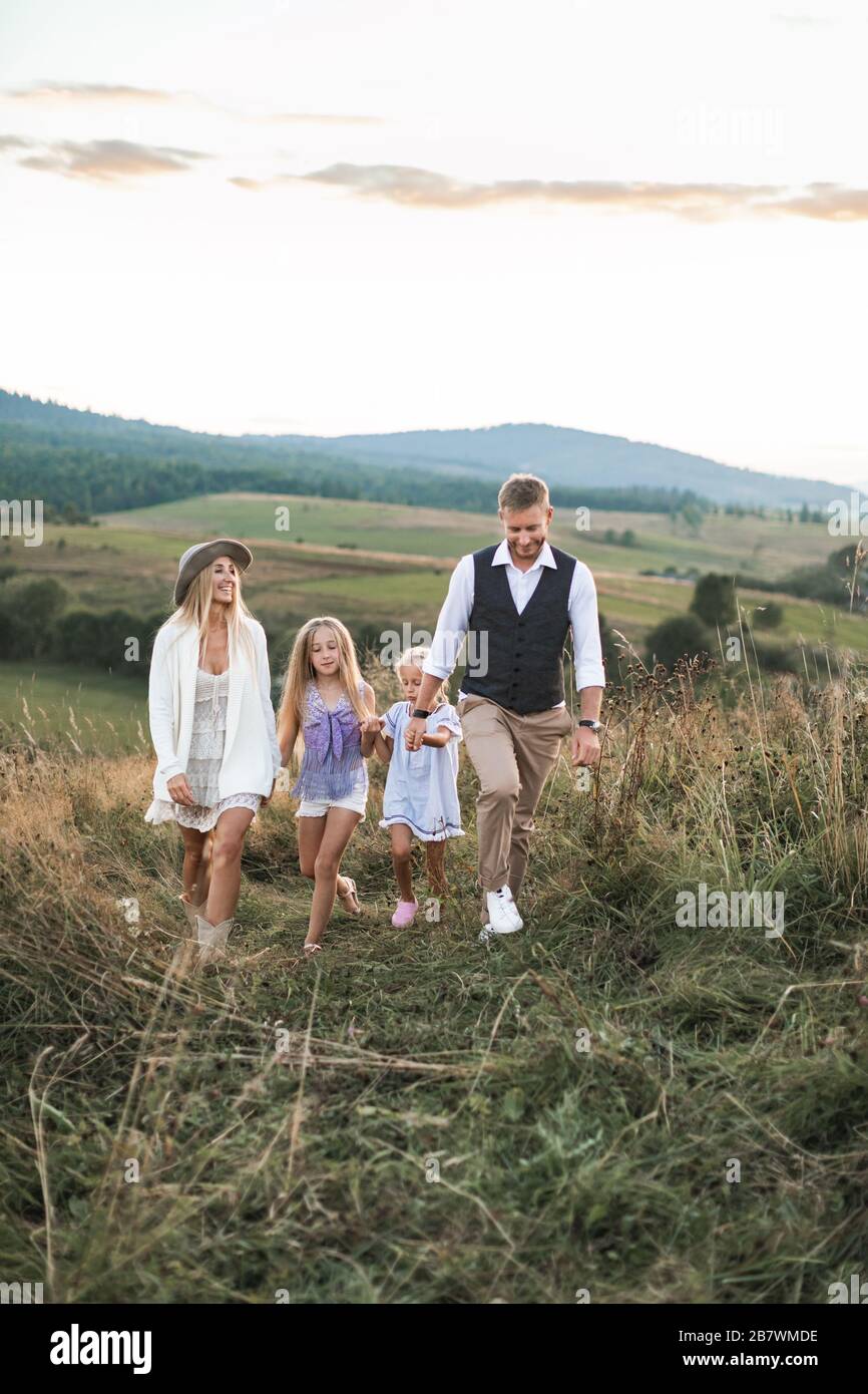 Feliz familia en ropa informal, madre, padre y dos hijas pequeñas en la naturaleza, caminar toethr con las manos, su mer puesta de sol en el campo salvaje en Foto de stock
