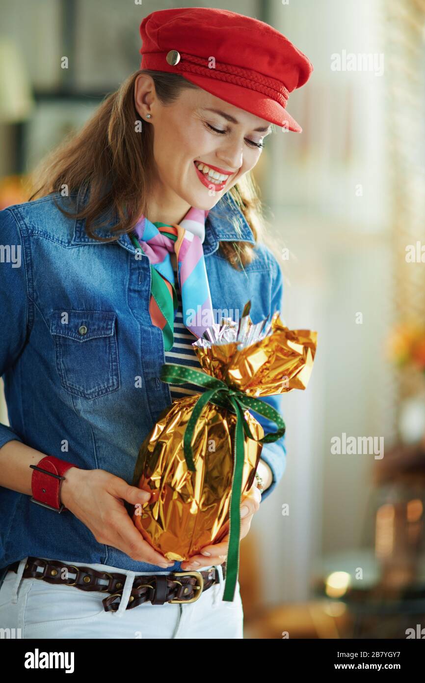 sonriente mujer moderna de edad media en una camisa de jeans y sombrero  rojo en la casa moderna en el soleado día de primavera con una celebración  envuelta en oro metalizado gran