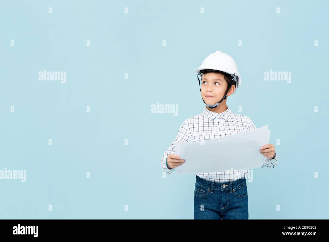 Sonriente joven asiático vestido como ingeniero con casco sosteniendo el plan de pensamiento y mirando hacia arriba en azul claro aislado estudio backgorund Foto de stock