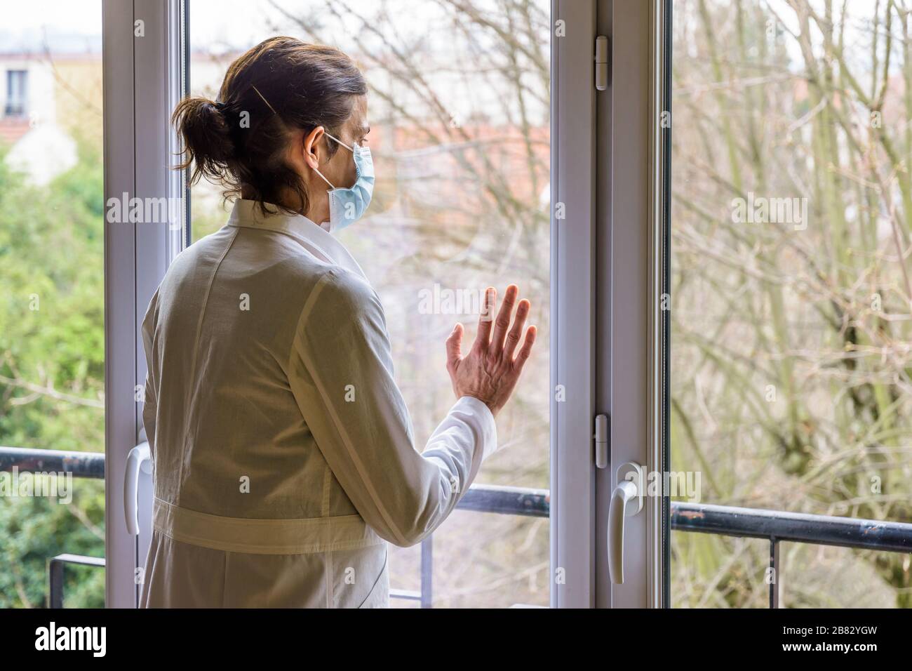 Una enfermera que lleva una máscara médica y un abrigo blanco, está de pie inactiva frente a una ventana cerrada, mirando hacia el espacio. Foto de stock