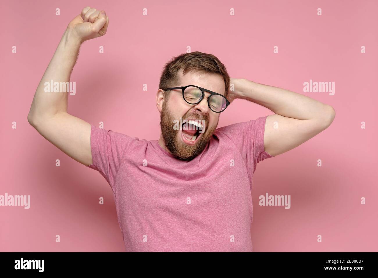 El hombre atractivo jovece con la boca abierta, cerrando los ojos y tirando de las manos, sobre un fondo rosa. Foto de stock