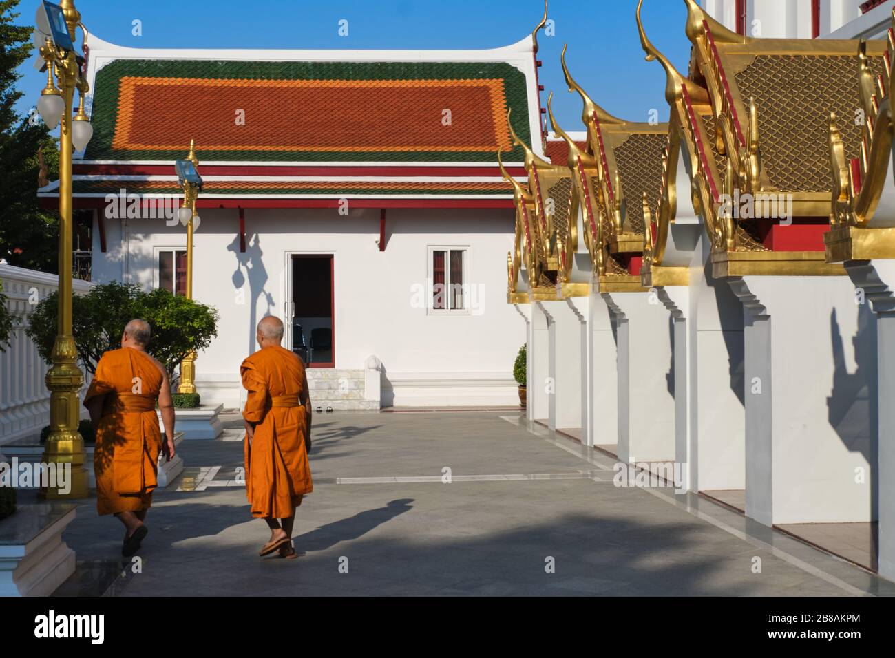 Dos monjes budistas de color naranja que caminan por los terrenos de Wat Ratchanadta, la base del icónico edificio Lohaprasad a la derecha; Bangkok, Tailandia Foto de stock