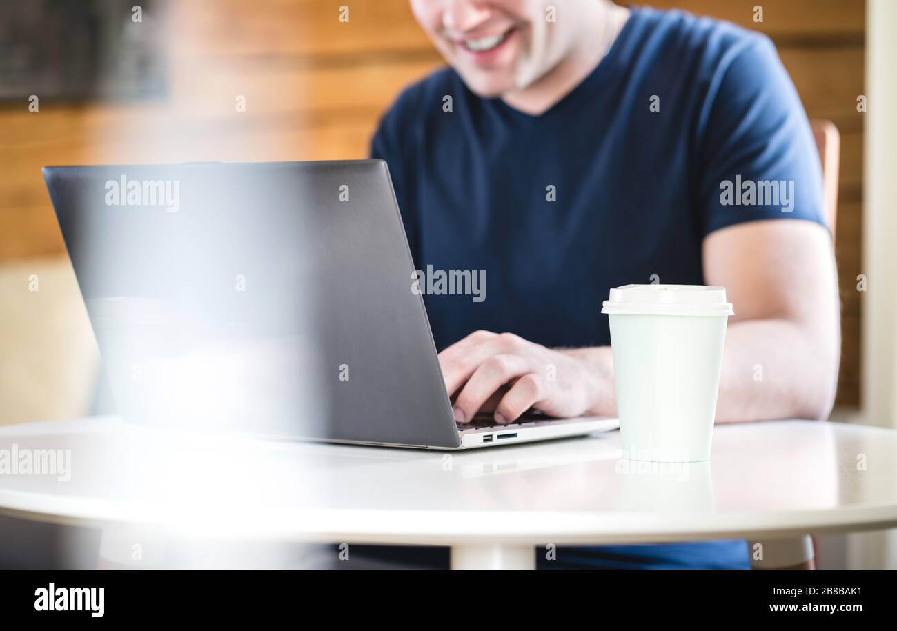 Hombre feliz usando el portátil con una taza de café para llevar sobre la mesa. Persona sonriente con ordenador en casa o en el café. Foto de stock