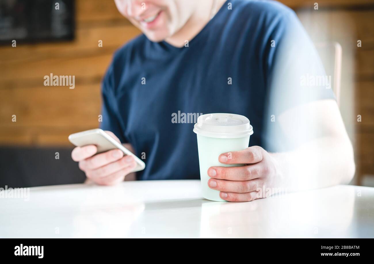 Feliz hombre usando smartphone en la cafetería o en casa. Hombre sonriente que sostiene el teléfono móvil y taza de café de papel. Mensajes de texto, compras en línea o navegar por Internet. Foto de stock