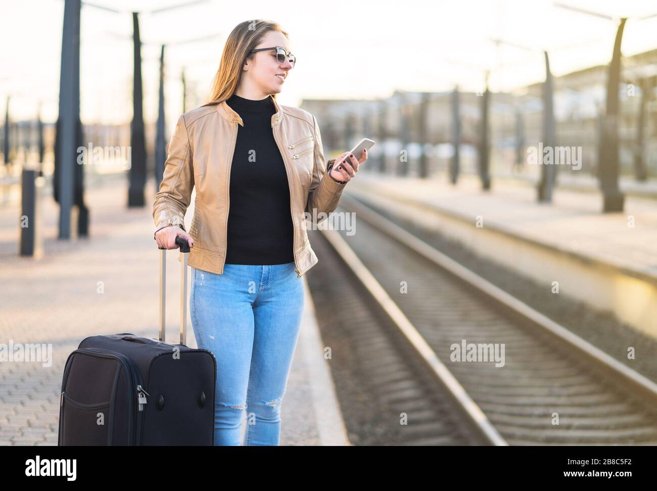 Esperando el tren. Mujer comprando boleto electrónico con teléfono móvil en plataforma. Pasajero en la estación con equipaje y maleta. Foto de stock