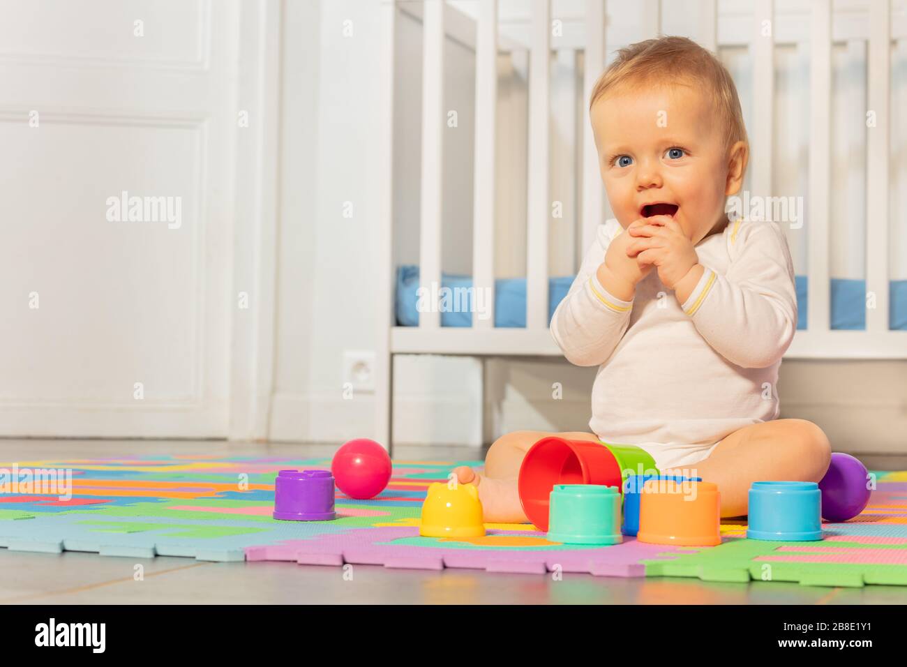 Hermoso bebé niño a la edad de niño, las manos cerca de la cara sentado frente a la cuna jugar con juguetes en la alfombra Foto de stock