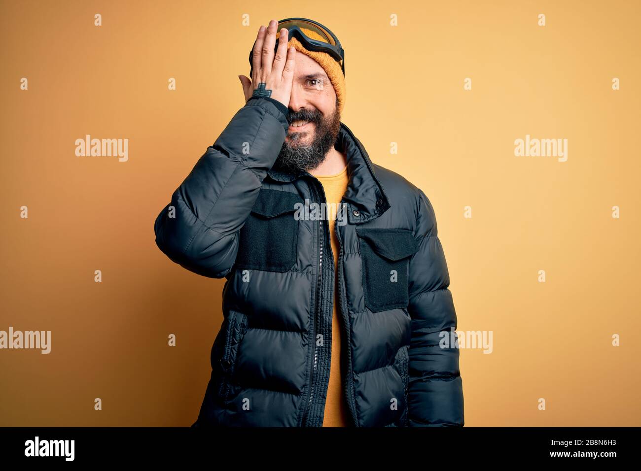 Un hombre calvo muy bonito esquiador con barba esquiando con ropa deportiva  de nieve y gafas de esquí que cubren un ojo con la mano, sonrisa segura en  la cara y sorpresa