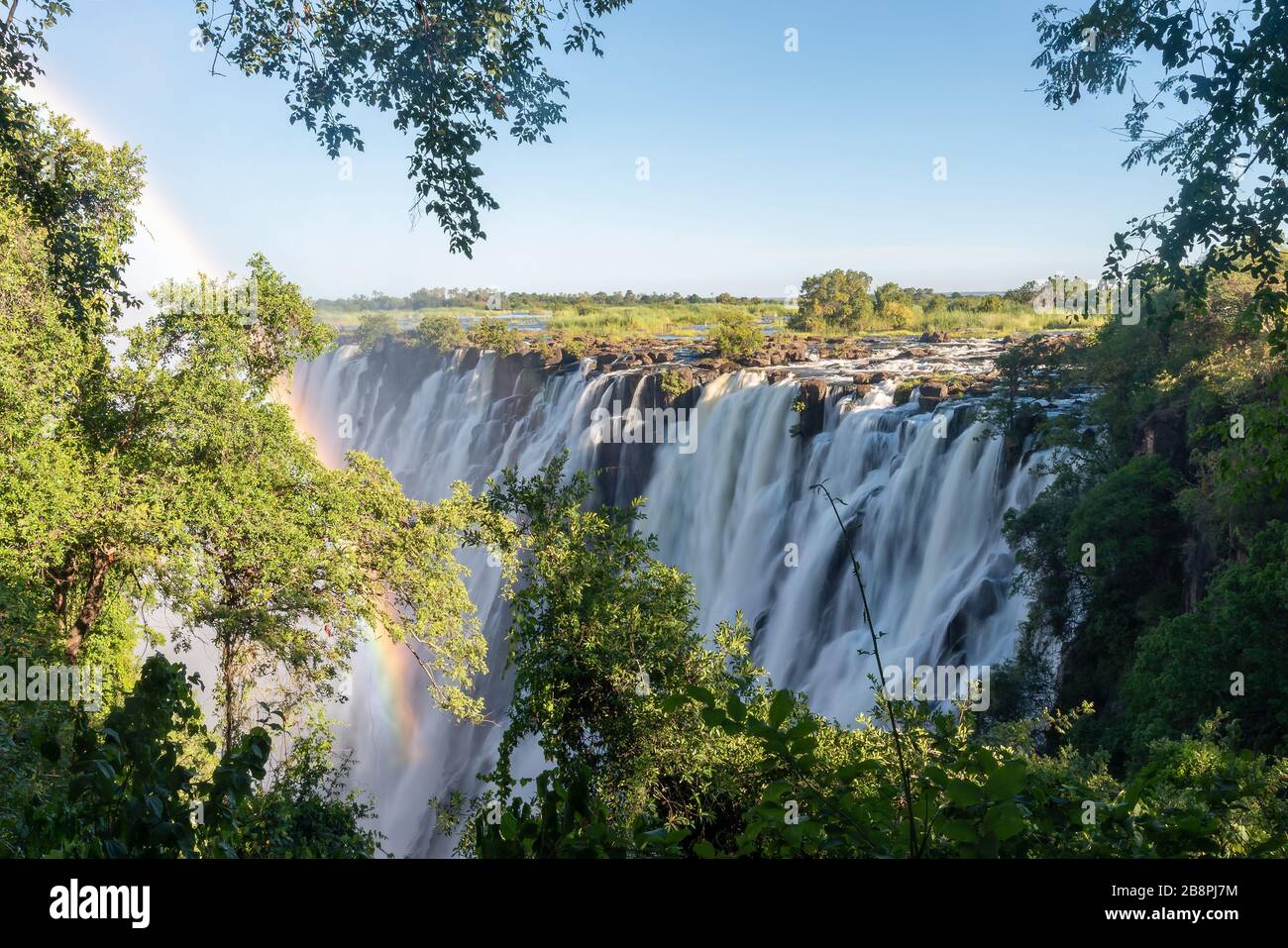 Cataratas Victoria, situadas en la frontera de Zimbabwe y Zambia. Arco iris y largo efecto de exposición del agua que fluye con vegetación y rocas. Foto de stock