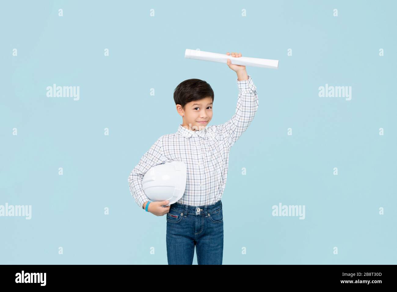 Lindo y sonriente joven asiático con casco que aspira a ser futuro ingeniero mostrando BluePrint aislado sobre fondo de estudio azul claro Foto de stock