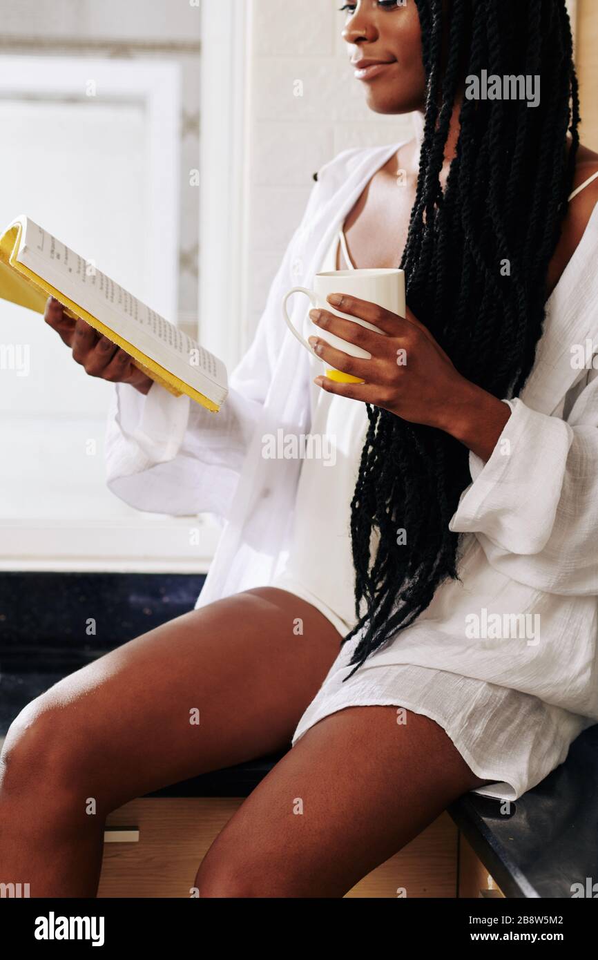 Hermosa mujer negra joven en pijama de lino bebiendo café de la mañana y leyendo un libro Foto de stock