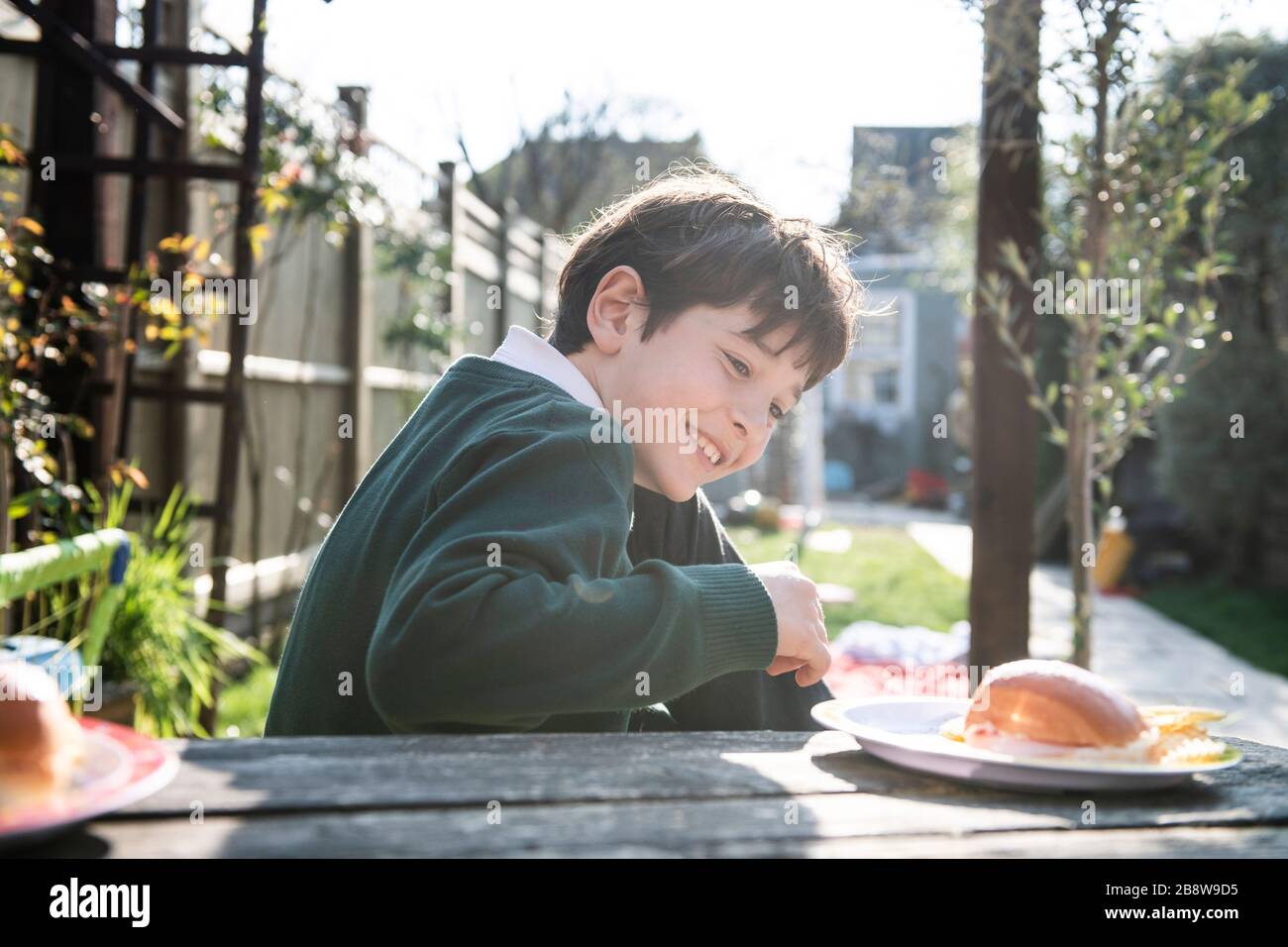 Niño sonriente con un Jersey verde sentado en una mesa en un jardín con un aperitivo. Foto de stock