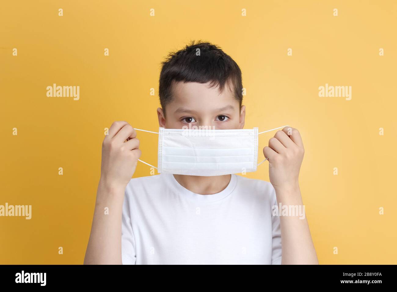 Niño caucásico con pelo oscuro en una camiseta blanca sosteniendo, poniendo una máscara médica sobre un fondo amarillo. Concepto de protección frente a infecciones virales Foto de stock