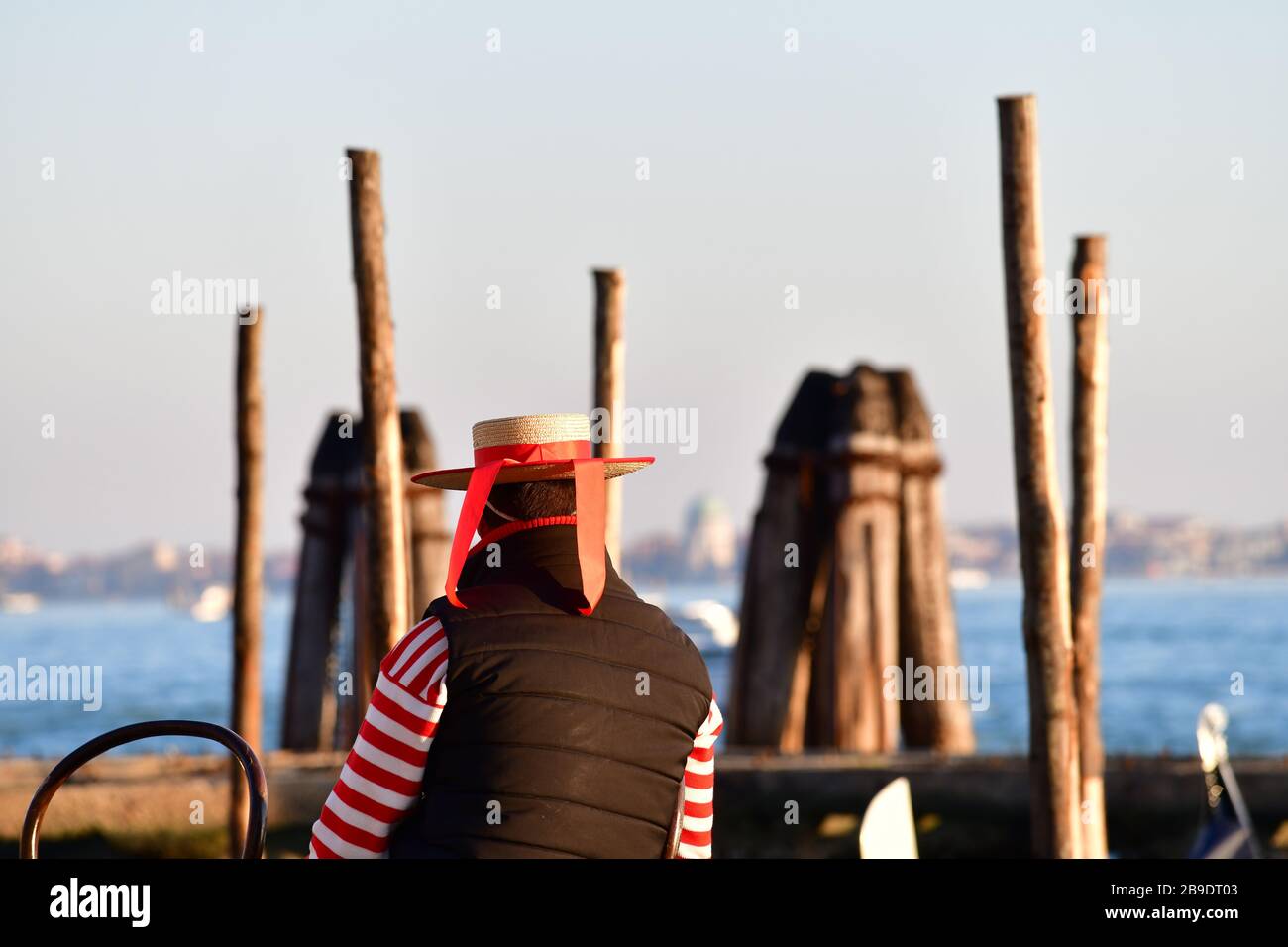 Gondolero con sombrero y camisa de rayas de color rojo-blanco que se reclinan con vistas a la laguna de Venecia desde una silla Foto de stock