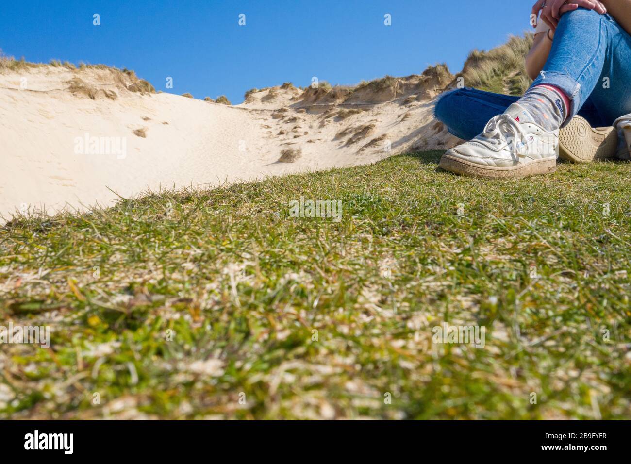 Una gran duna de arena con las piernas de las chicas en primer plano sobre hierba con zapatillas y jeans Foto de stock