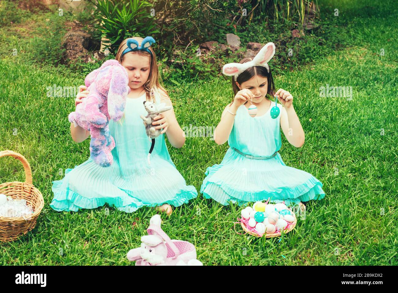 Niños pequeños y lindos que usan orejas de conejito en el día de Pascua sentado en el jardín y jugando con huevos pintados y conejito de Pascua y juguetes de bibby. Foto de stock