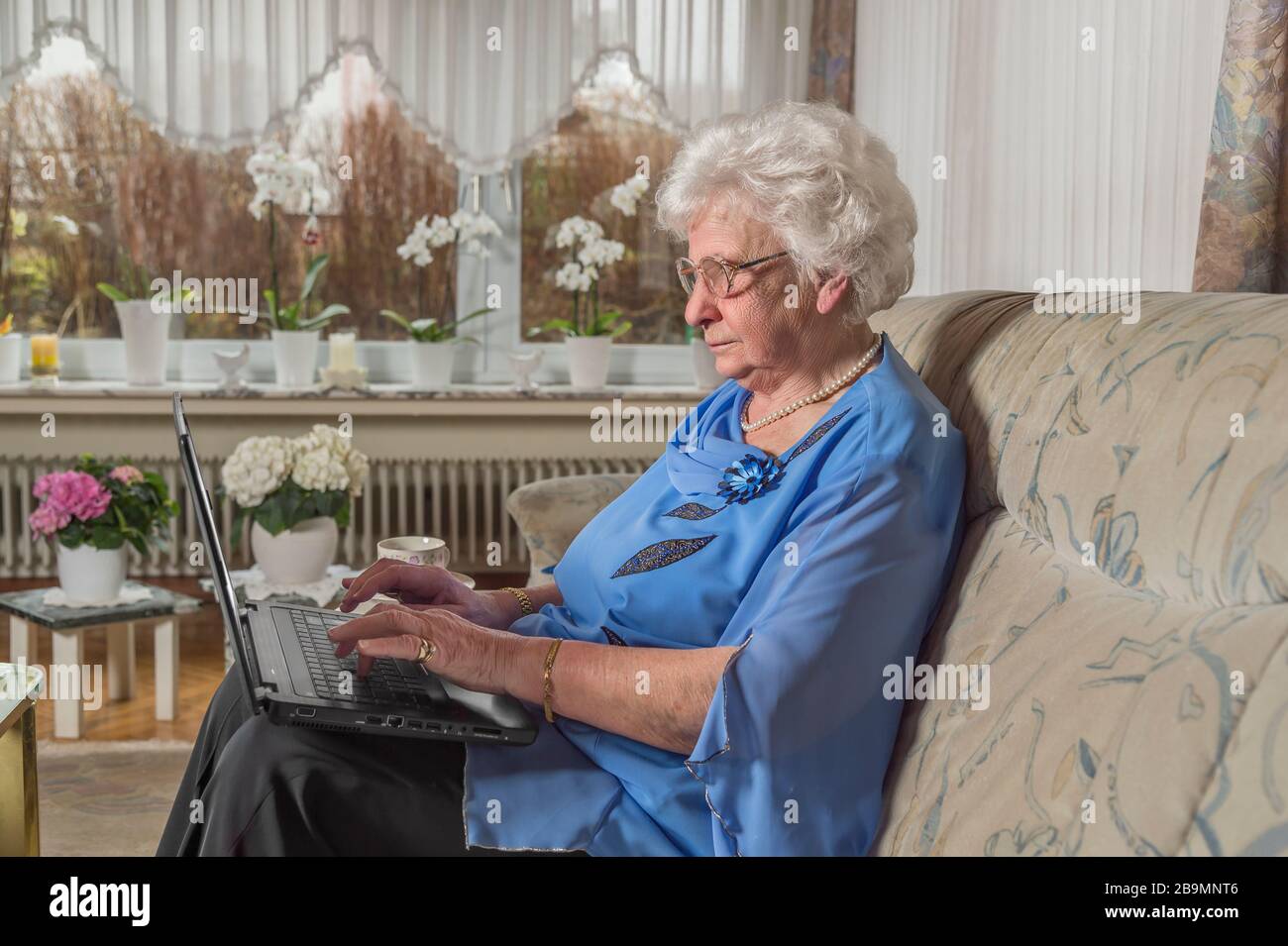 mujer de 80 años sentada sola en casa. Pertenece al grupo de riesgo de Covid 19. La mujer mayor escribe en el portátil contra el aislamiento con su familia Foto de stock