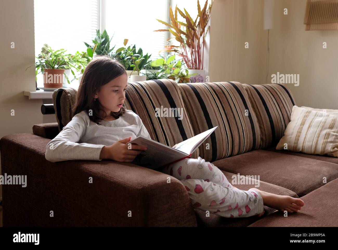 La niña está leyendo un libro en el sofá. Aprendizaje a distancia. Foto de stock