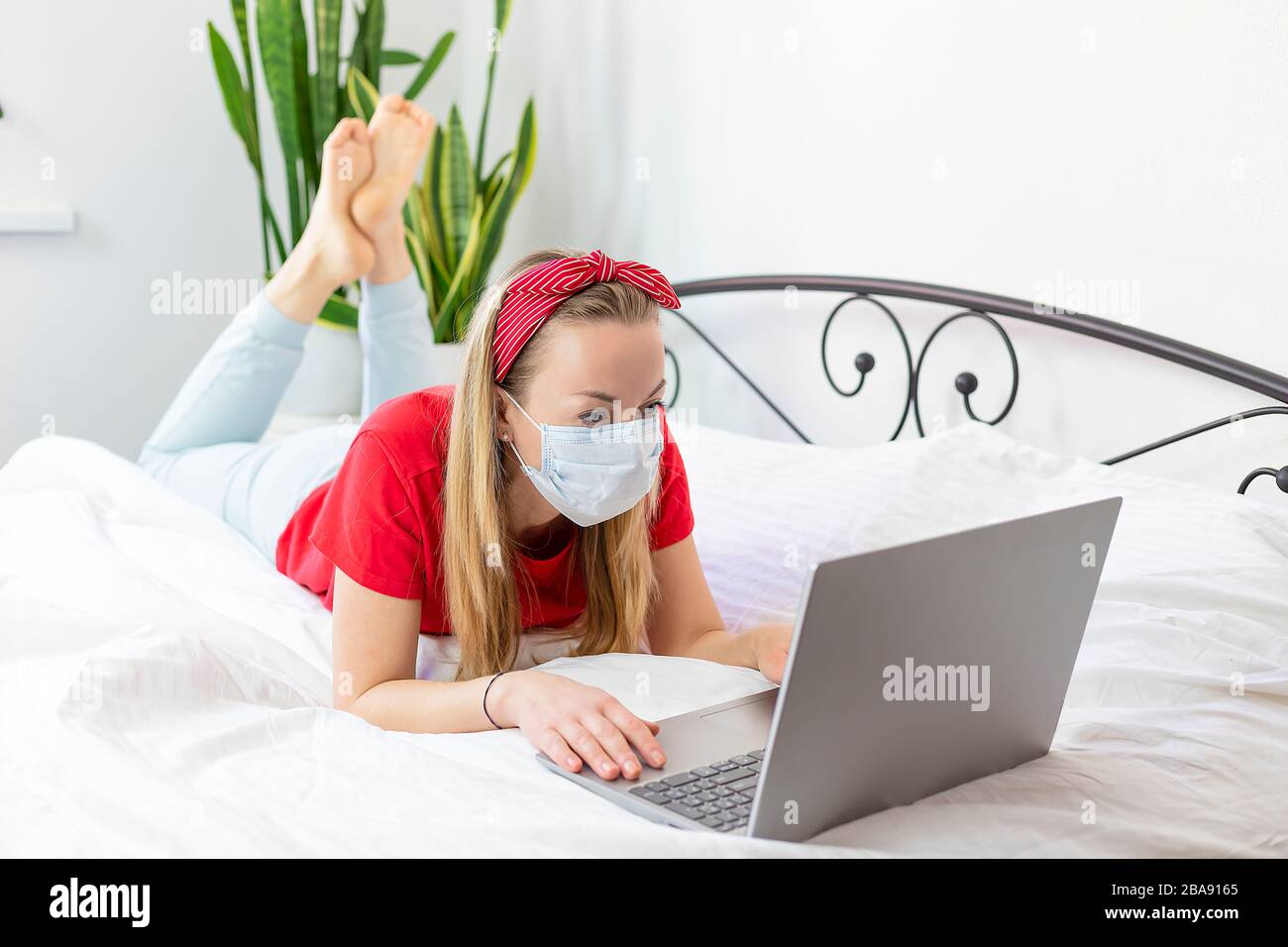 mujer joven enferma con una máscara médica y una camiseta roja y pantalones cómodos se encuentra en una cama blanca con un ordenador portátil. trabaja desde casa debido al coro Foto de stock