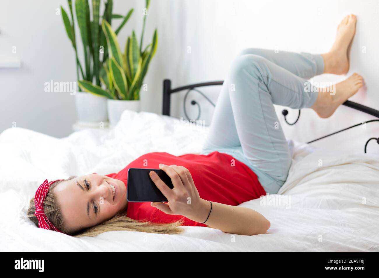 Mujer joven sonriente con pelo rubio en una camiseta roja y pantalones cómodos se encuentra en una cama blanca con un smartphone. Trabaja desde casa, escribe y lee Foto de stock