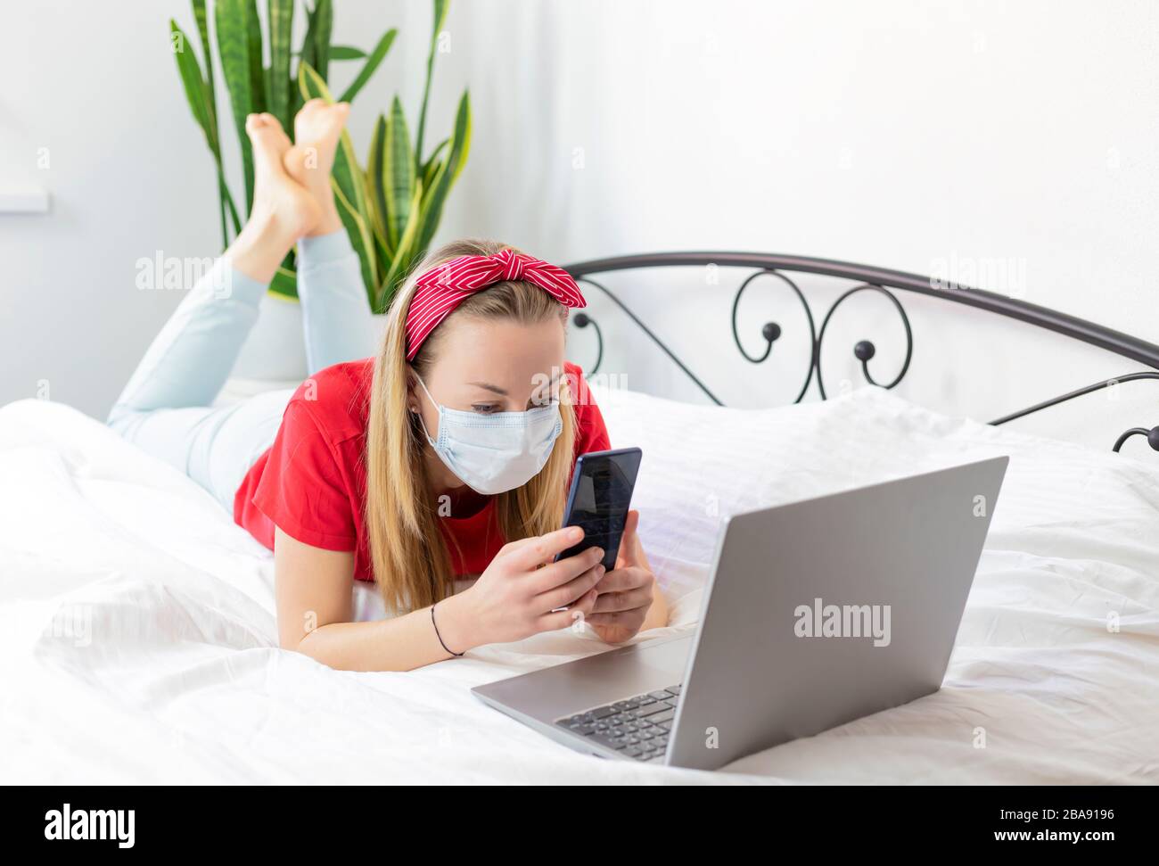 una joven enferma con una máscara médica y una camiseta roja y pantalones cómodos se encuentra en una cama blanca con un ordenador portátil y un teléfono. trabaja desde casa porque Foto de stock