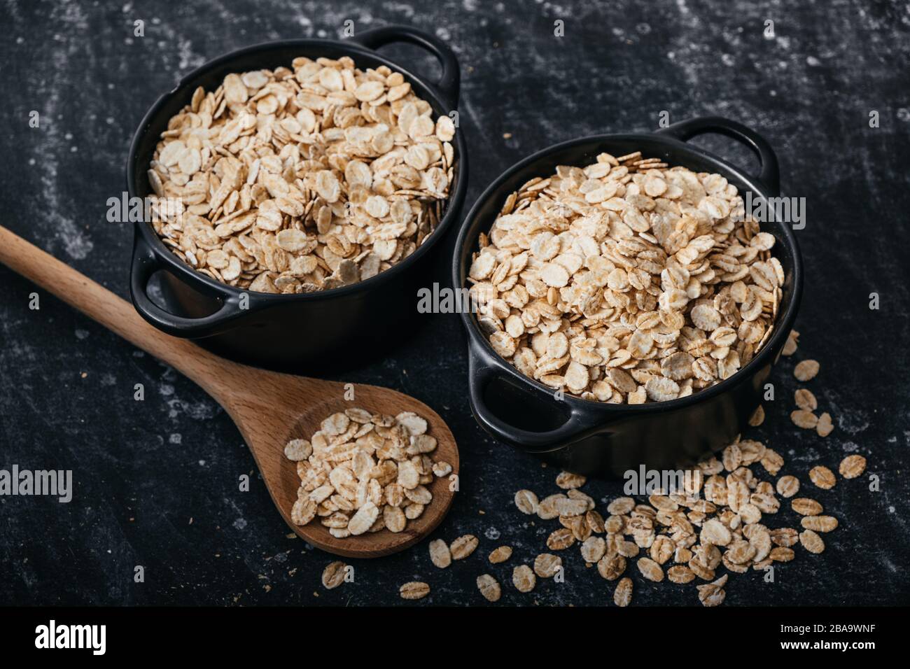 Vista superior de dos cuencos negros y cuchara con avena cruda sobre fondo negro. Desayuno con fibras para una dieta equilibrada. Foto de avena Foto de stock