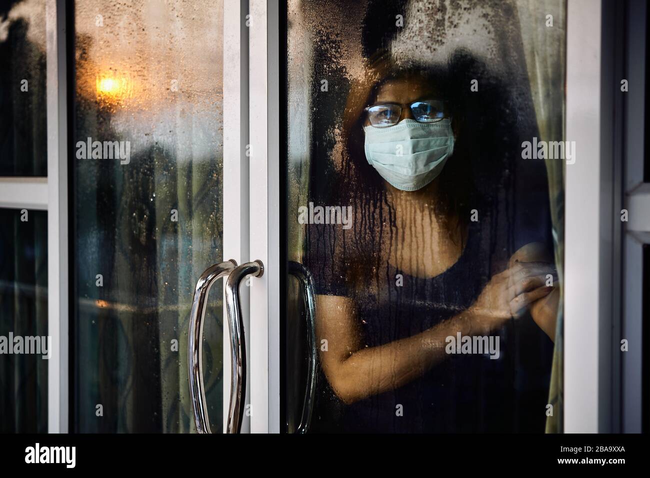 Mujer mirando a través de la ventana húmeda durante la estancia en casa mientras COVID-19 Foto de stock