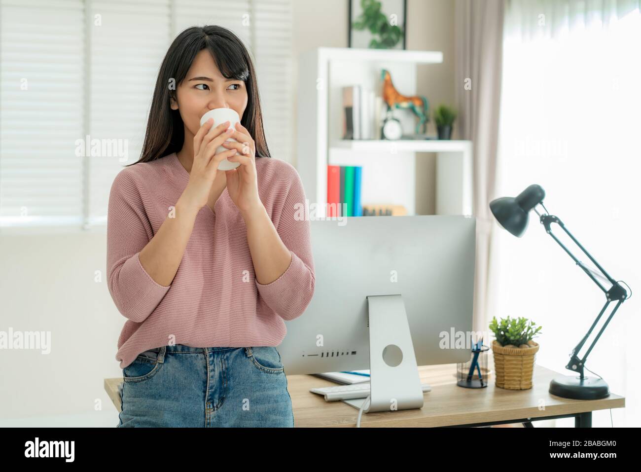 Una hermosa mujer joven asiática trabaja desde casa trabajando en la computadora y bebiendo café mientras se relaja de su trabajo de pie en la sala de estar en casa. Di social Foto de stock