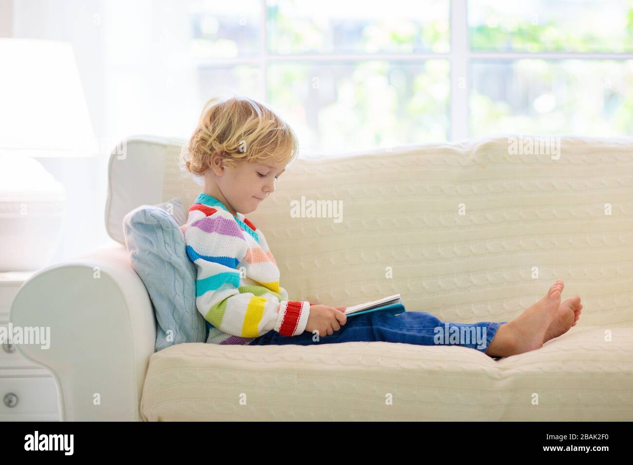 Libro de lectura infantil. Leer libros para niños. Chico sentado en el sillón blanco, en la soleada sala de estar viendo imágenes de libro de cuentos. Kid haciendo los deberes para el Foto de stock