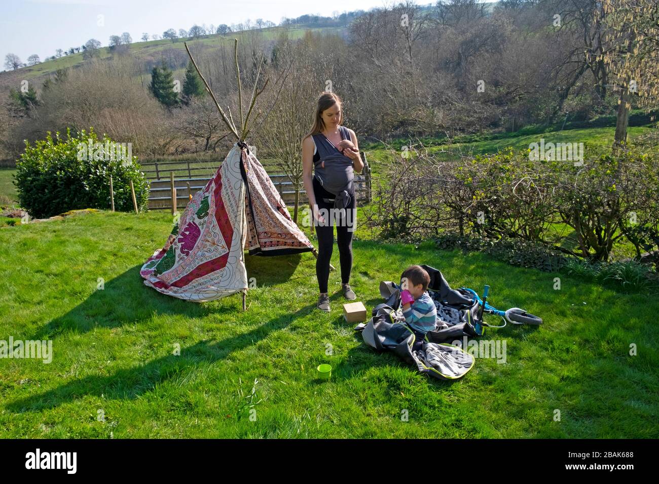 Madre de pie con un portador de bebé y un niño por tipi tienda jugando en un jardín de campo durante Covid-19 brote primavera 2020 Gales Reino Unido KATHY DEWITT Foto de stock