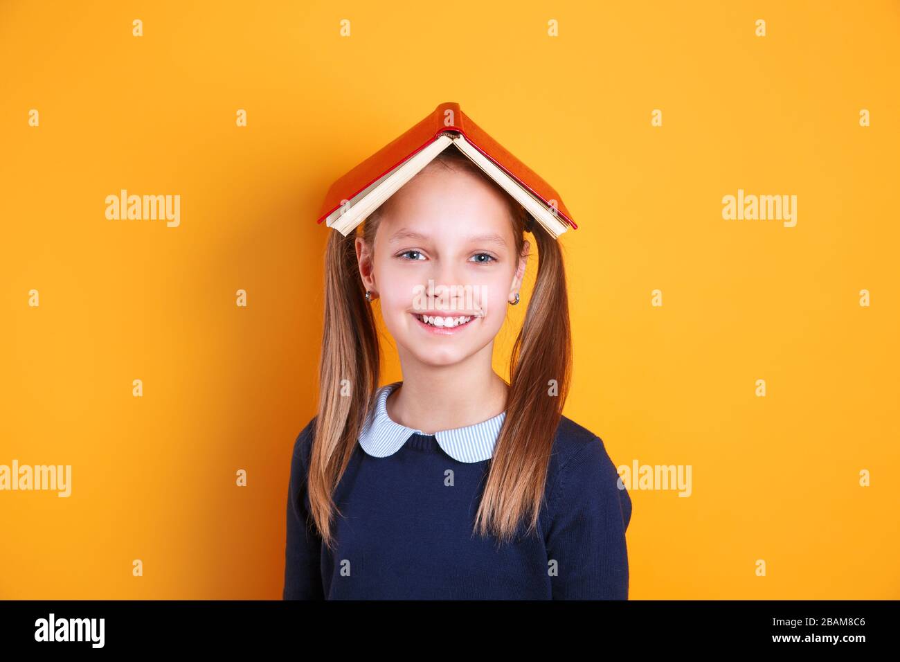 cute niña de la escuela sosteniendo el libro sobre la cabeza como techo sobre fondo amarillo Foto de stock