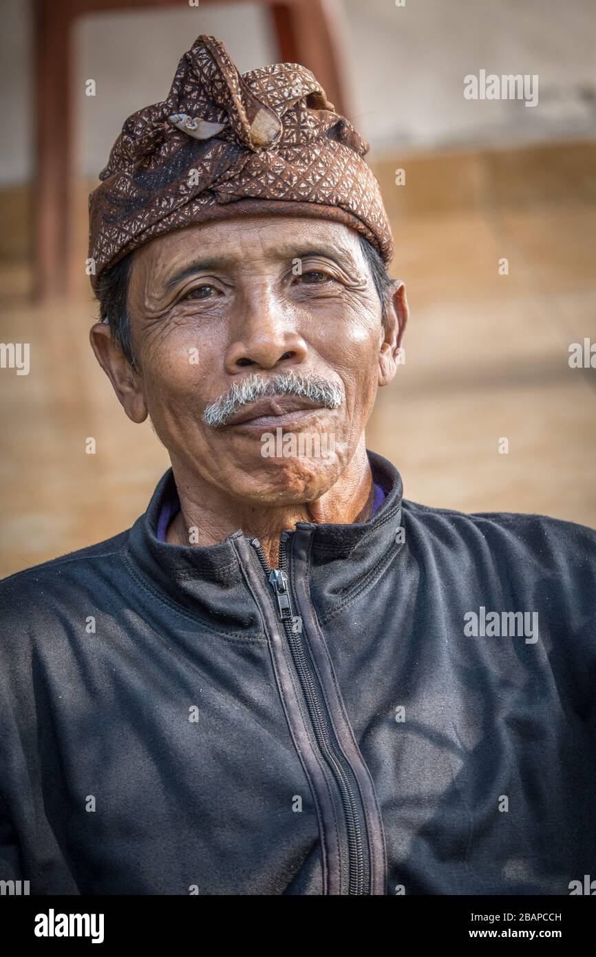 Retrato de un anciano que comprueba las entradas en el templo Pura Gunung Kawi, con un tocado de batik marrón, chaqueta de cuero negro con ojos y bigote. Foto de stock