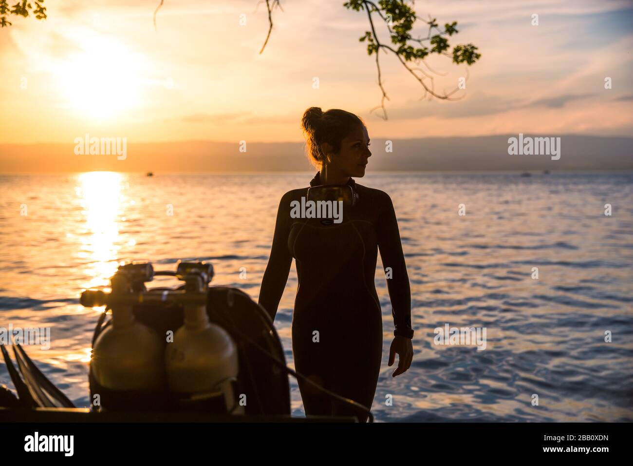 Instructor femenino de buceo usando un traje húmedo de pie junto a un tanque doble en Sunset Foto de stock