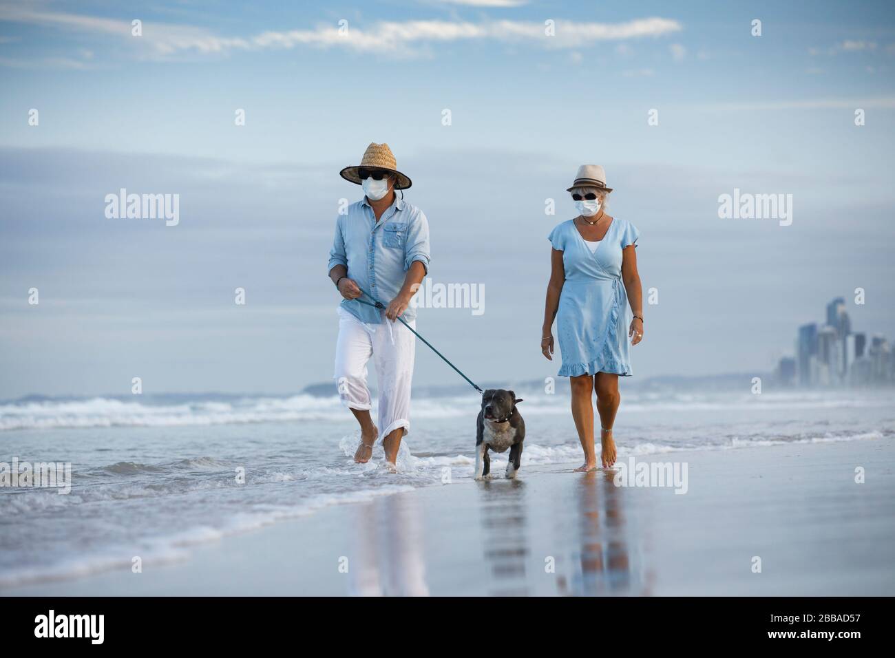 una pareja con máscaras médicas caminando por un perro azul stafford en la playa. un perro caminante o permanecer activo en medio de la pandemia del coronavirus Foto de stock