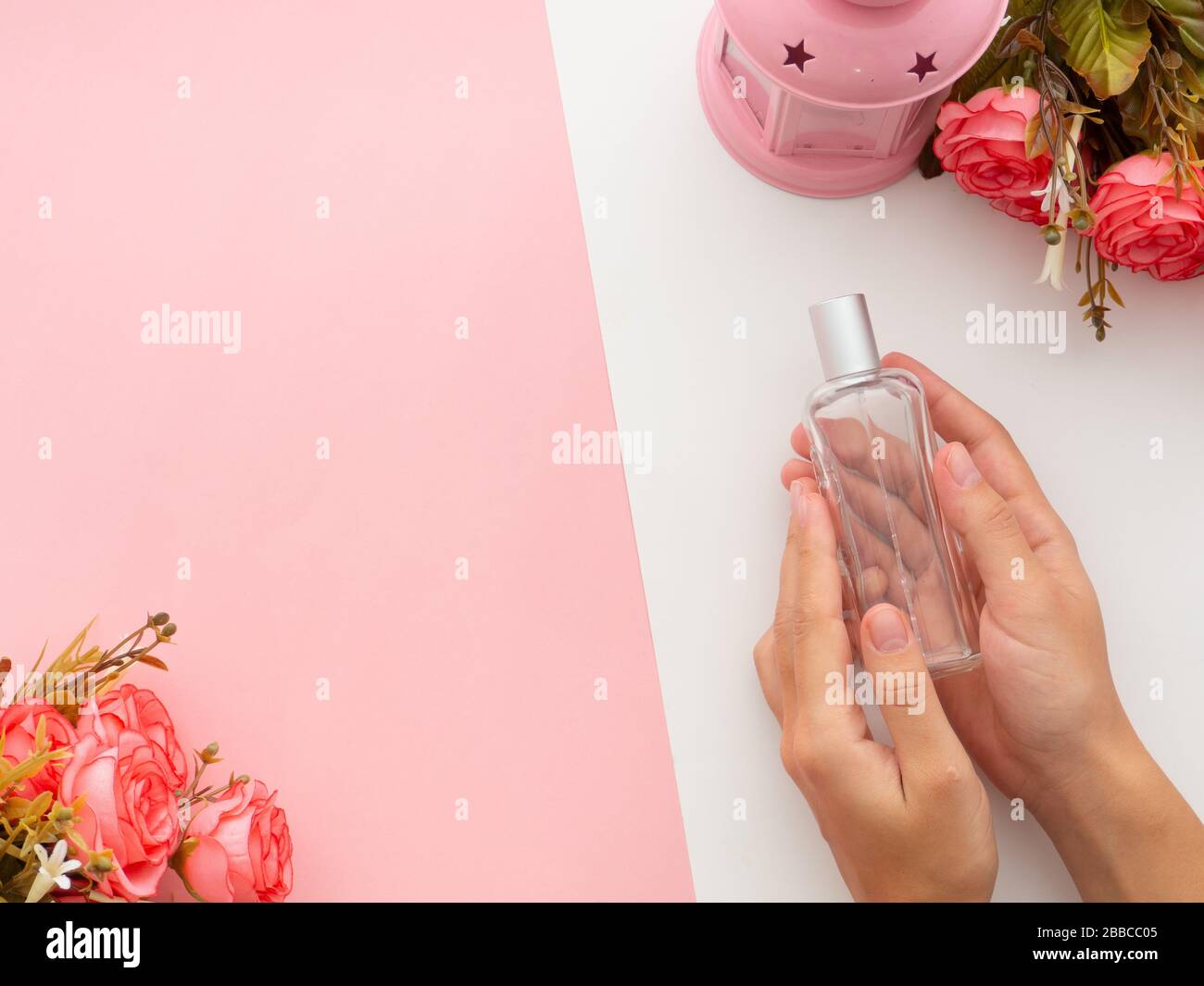 Concepto de perfumería y aroma floral. Manos sosteniendo una elegante  botella de perfume con flores rosas alrededor y una lámpara de luz sobre  fondo rosa blanco. Top vie Fotografía de stock -