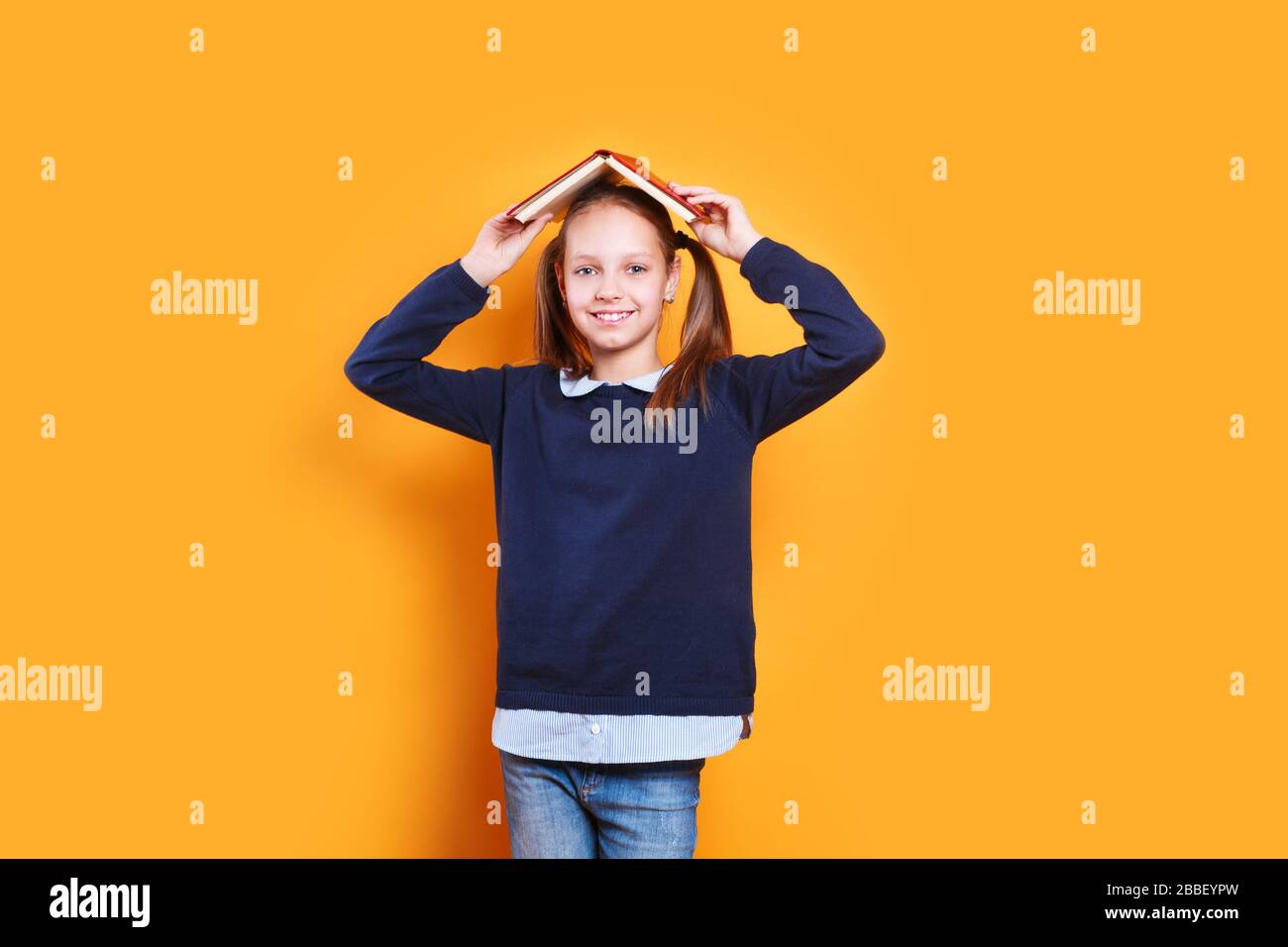 cute niña de la escuela sosteniendo el libro sobre la cabeza como techo sobre fondo amarillo Foto de stock