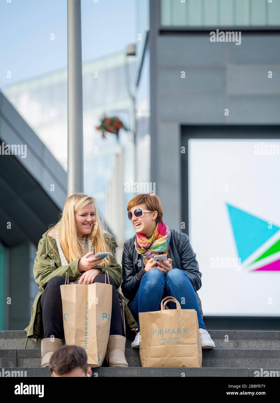 Dos amigos toman un descanso sentado y comparten una broma mientras compran en el centro comercial Westfield, Stratford. Completo con bolsas de compras y teléfonos. Foto de stock