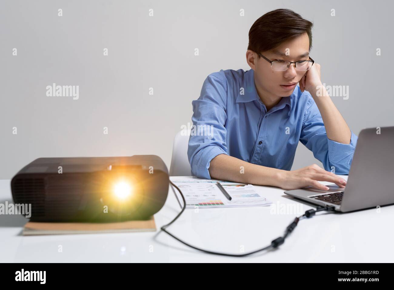 Un joven analista asiático en gafas sentado en el escritorio con proyector y utilizando el portátil mientras trabaja en la presentación Foto de stock