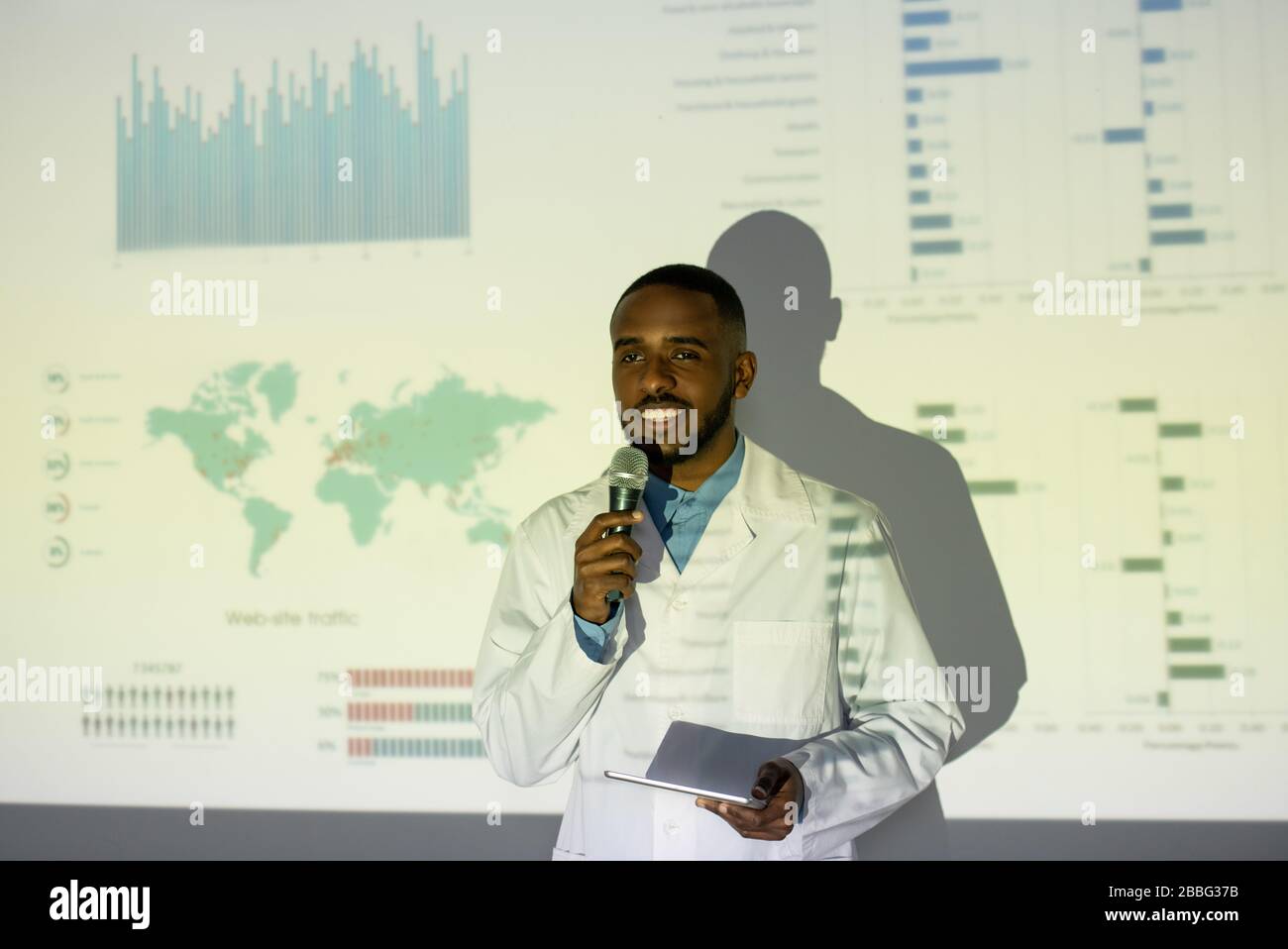 Sonriente joven científico médico afro-americano en el abrigo de laboratorio de pie contra la pantalla de proyección y ofrecer la medicación coronavirus en la convención Foto de stock