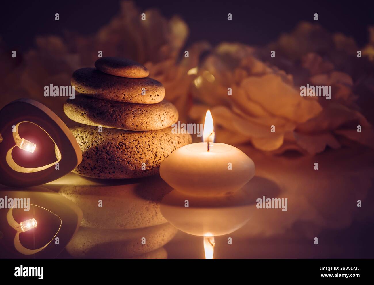 Piedras planas redondas zen apiladas en el spa con llamas de velas y reflejo en la oscuridad, flores borrosas pétalos sobre el fondo. Foto de stock