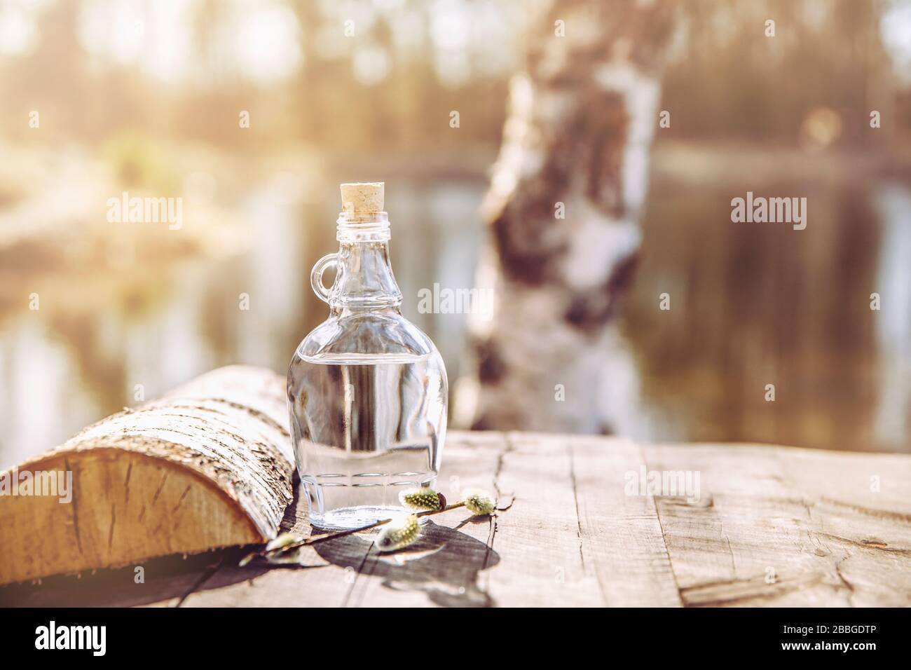 Jugo de abedul fresco filtrado savia en botella de vidrio transparente sobre mesa rústica amarilla, al aire libre en primavera, troncos de abedul en el fondo. Foto de stock