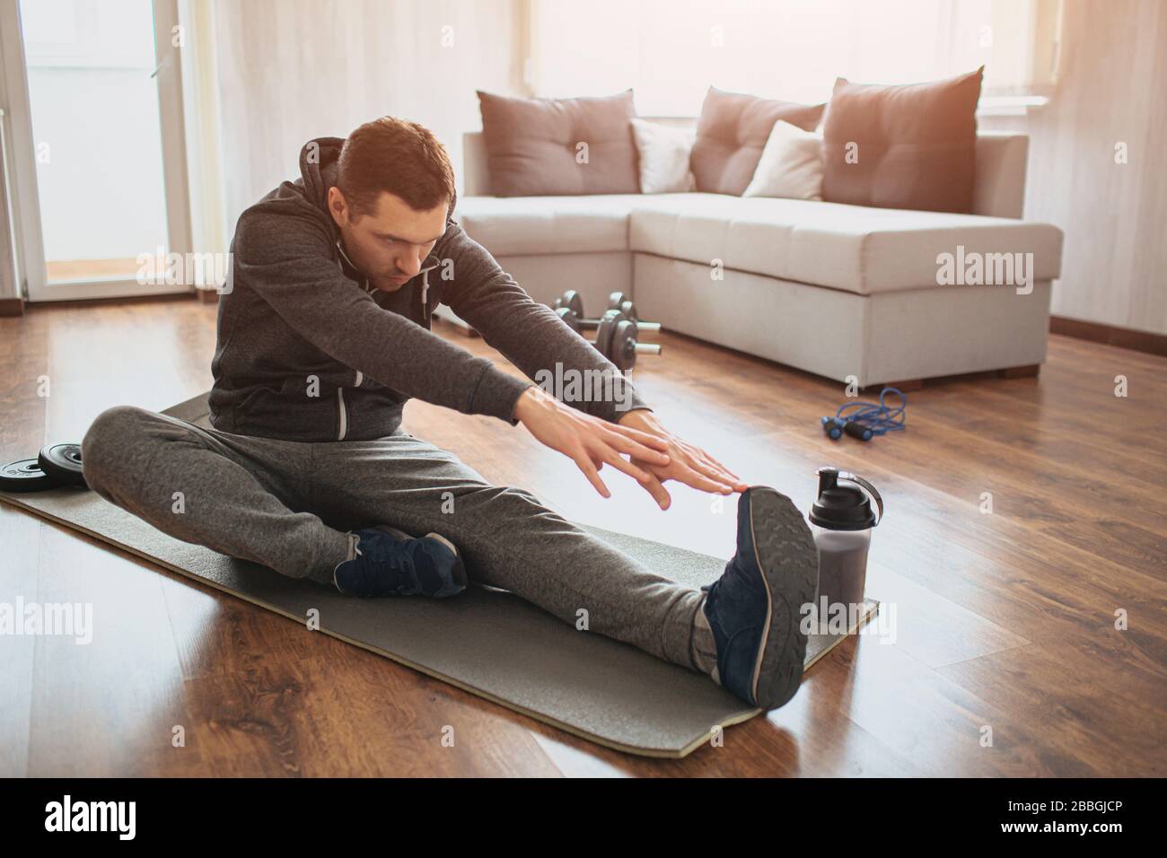 El joven hombre común va a practicar deporte en casa. Freshman o principiante estirando con las manos de remolque a una pierna. Excercising solo en apartamento vacío. Inténtelo a. Foto de stock