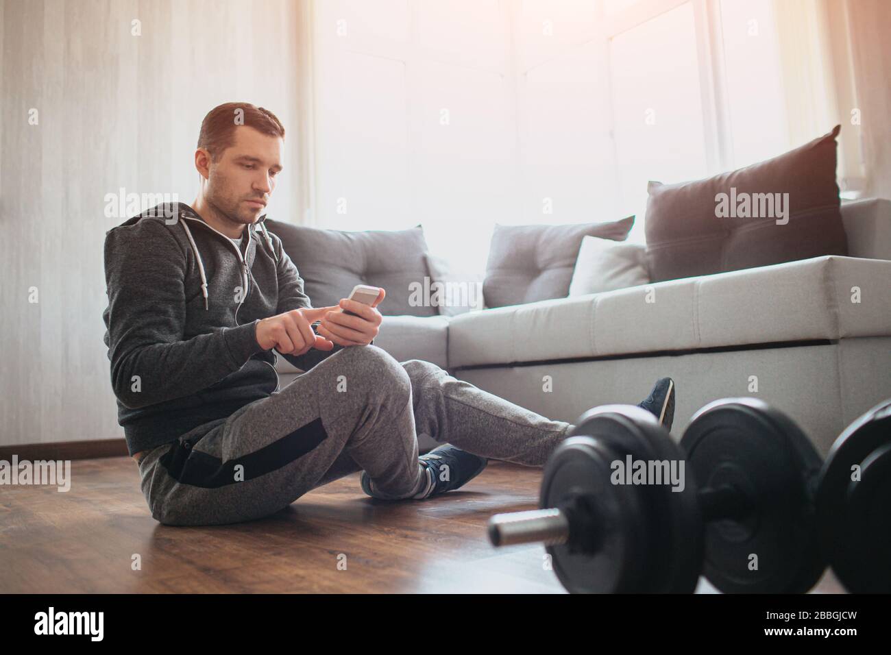 El joven hombre común va a practicar deporte en casa. Tiene descanso con sentarse en el suelo y utilizar el smartphone. Refrescar en el entrenamiento relajándose después del entrenamiento. Solo en Foto de stock