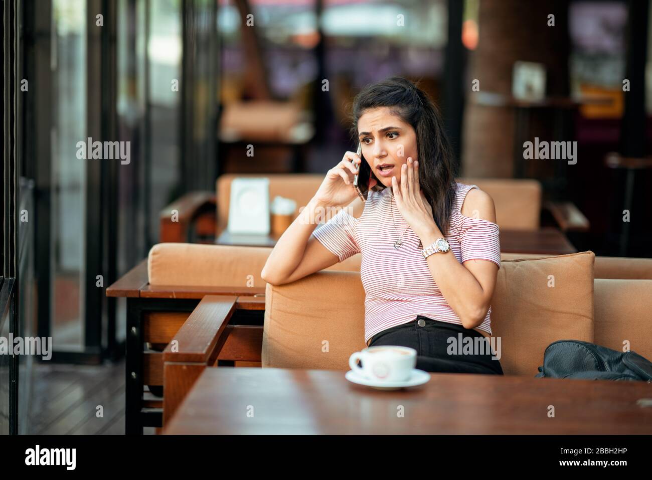 Una mujer joven habla con el teléfono y tiene una cara sorprendida y preocupada en un café elegante Foto de stock