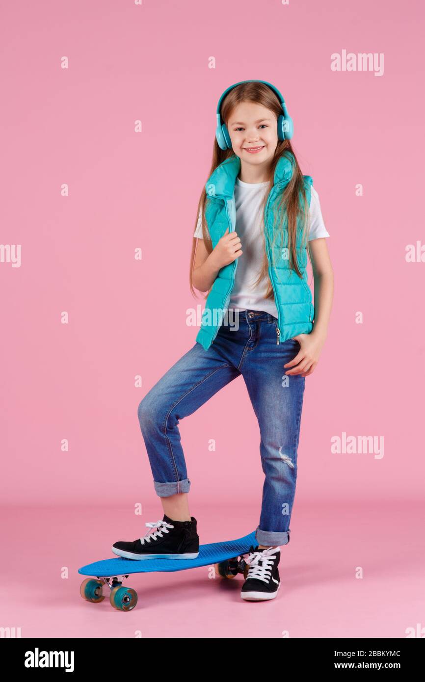Hermoso niño rubio niña de 7-10 años en chaqueta posando aislado en color rosa pastel fondo niños retrato. Concepto de estilo de vida infantil. Crear espacio de copia. Mantén el skateboard azul Foto de stock